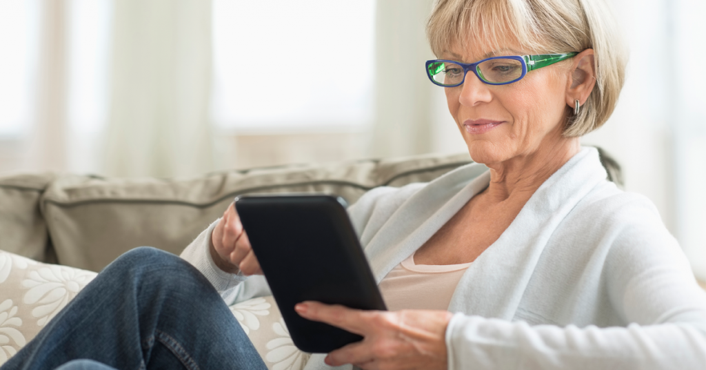 Older woman wearing green glasses sitting on a couch looking at a tablet