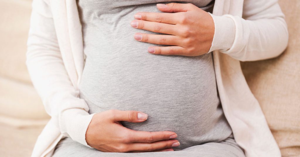 Pregnant belly in a grey tshirt with the mother's hands at the top and bottom